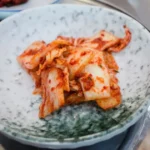 A bowl of homemade kimchi with a chopstick, representing the daily kimchi benefits for gut health and its probiotic power.